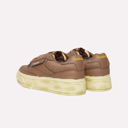 A pair of Reebok Club C LTD Shoes in brown with yellow logo accents and thick off-white soles, featuring a vintage-inspired, slightly worn look—shown from both back and side angles.