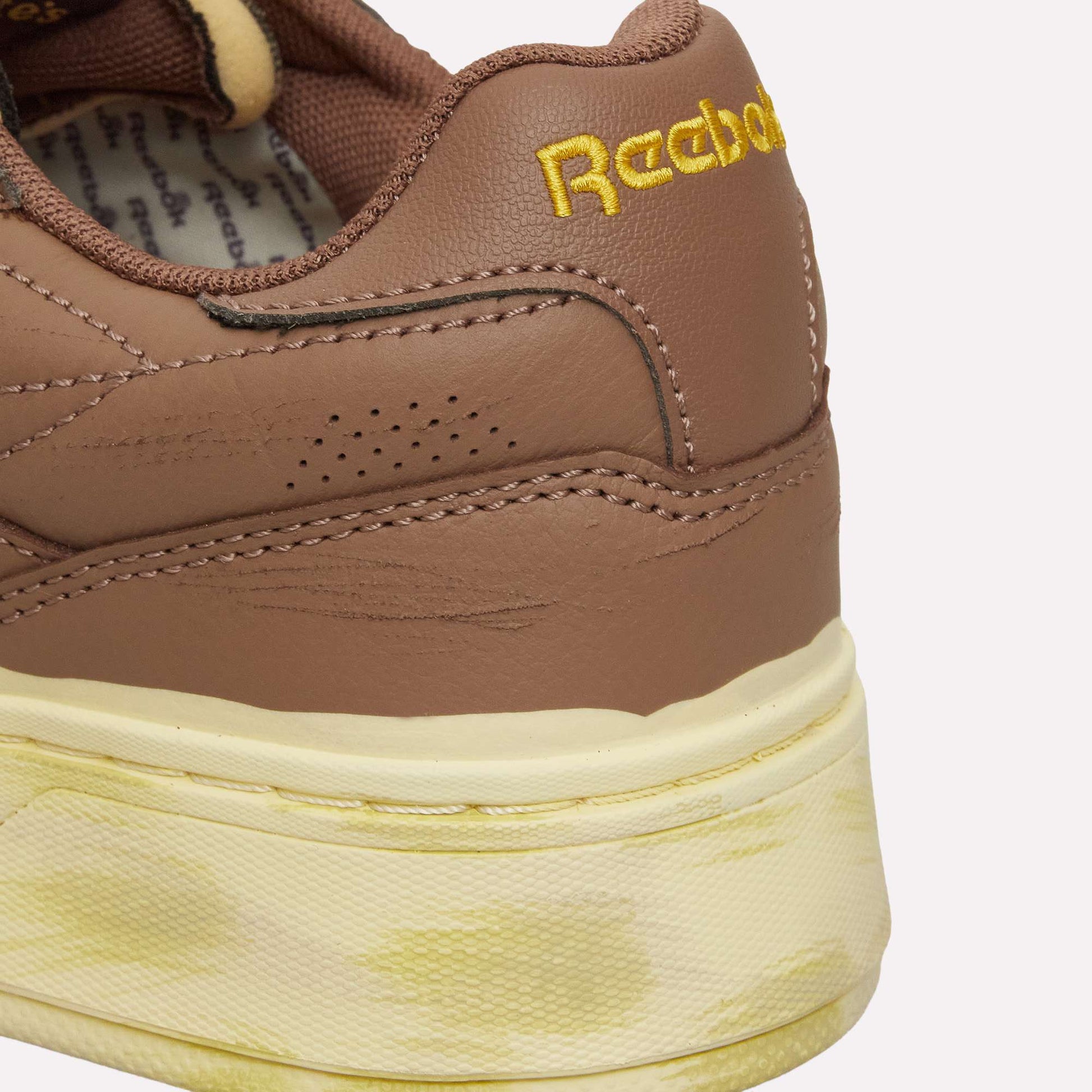 Close-up of the back of a brown Reebok Club C LTD shoe with yellow embroidered branding and a cream-colored midsole displaying yellowish stains, highlighting the timeless appeal of this classic from Reebok.