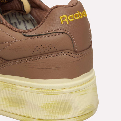 Close-up of the back of a brown Reebok Club C LTD shoe with yellow embroidered branding and a cream-colored midsole displaying yellowish stains, highlighting the timeless appeal of this classic from Reebok.