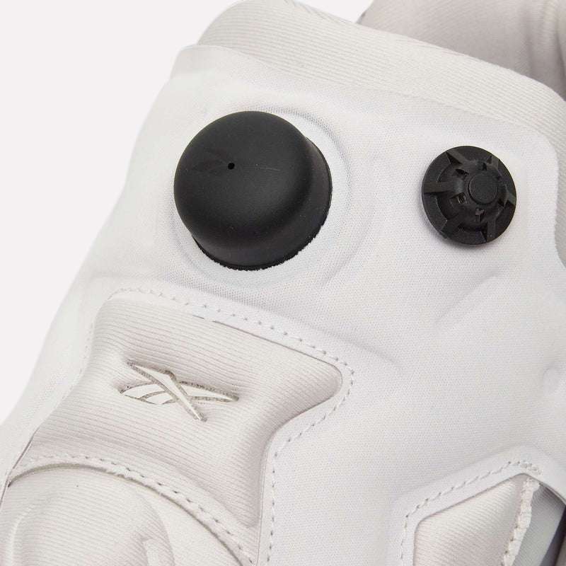 Close-up of a white Reebok Instapump Sliver Shoe with a black circular air pump and smaller black valve on the tongue. The Reebok logo is stitched in white on the side, set against a light blue background.
