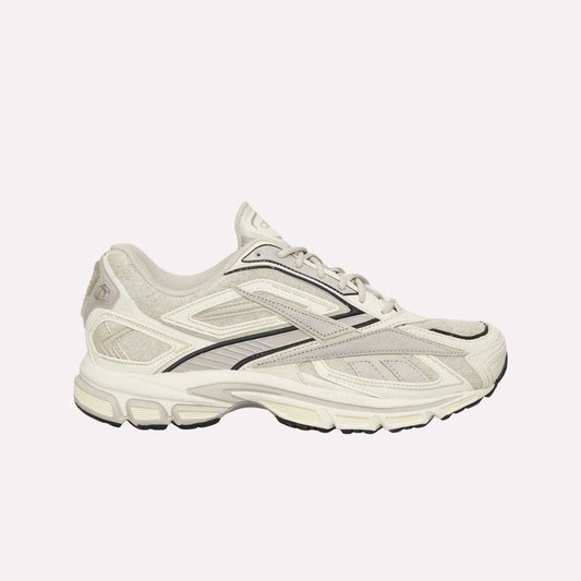 The Reebok Premier Road Ultra Shoes feature beige and white uppers with black accents, mesh panels, and a textured sole with a dual-density midsole, shown against a light blue background.
