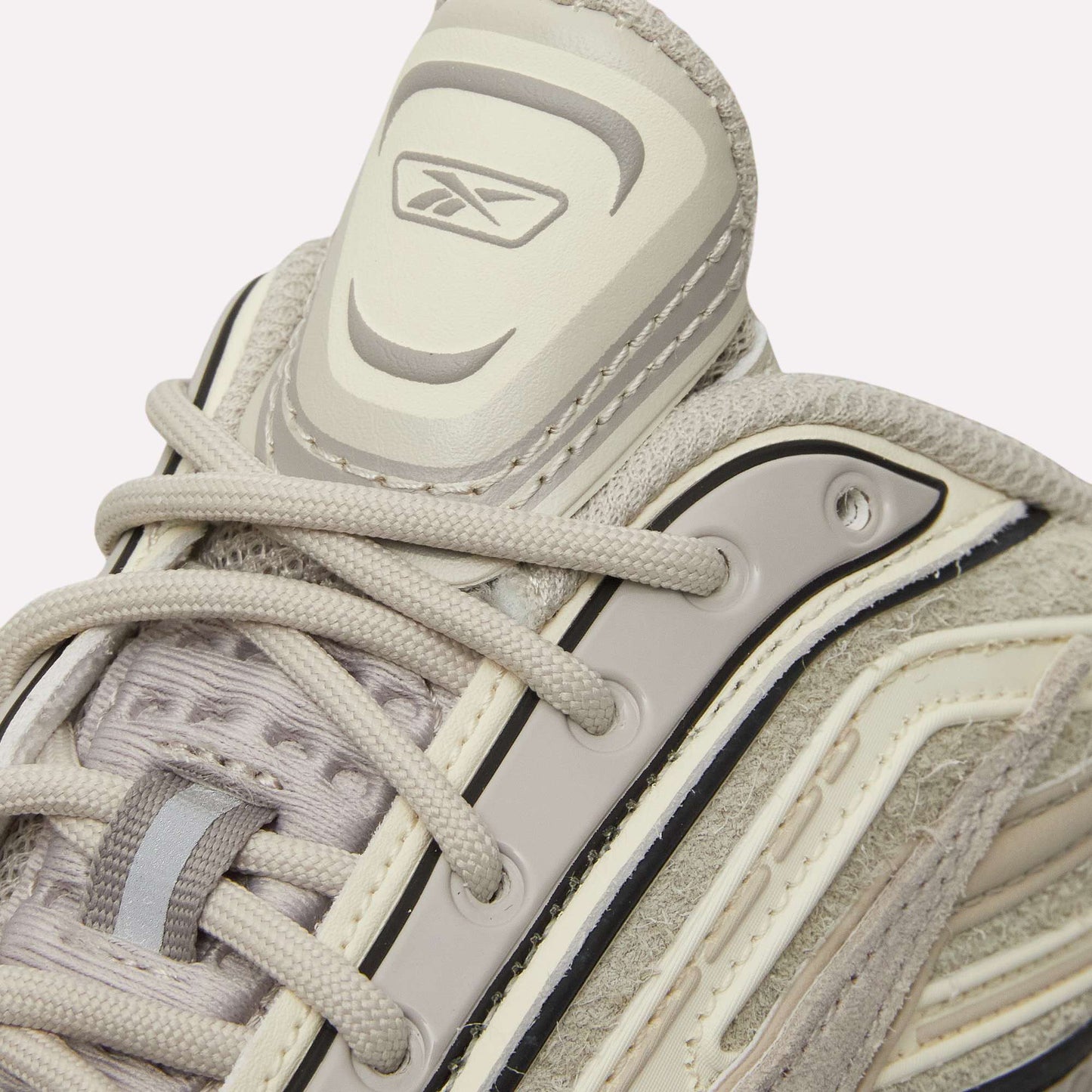 Close-up of Reebok Premier Road Ultra Shoes in beige and light gray with detailed stitching, dual-density midsole, laces, and Reebok logo on the padded tongue, against a light blue background.