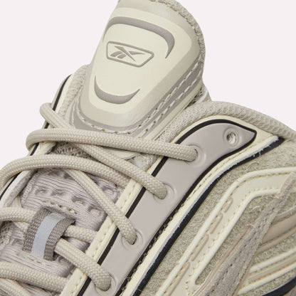 Close-up of Reebok Premier Road Ultra Shoes in beige and light gray with detailed stitching, dual-density midsole, laces, and Reebok logo on the padded tongue, against a light blue background.