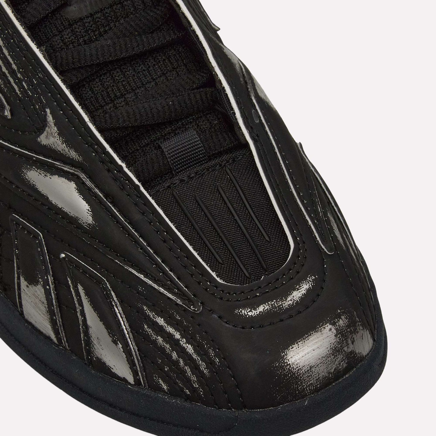 Close-up of the front and side of Reebok Hammer Pro LTD Shoes in black, featuring a shiny, textured faux leather upper with black laces and detailed stitched and grooved designs on the material.