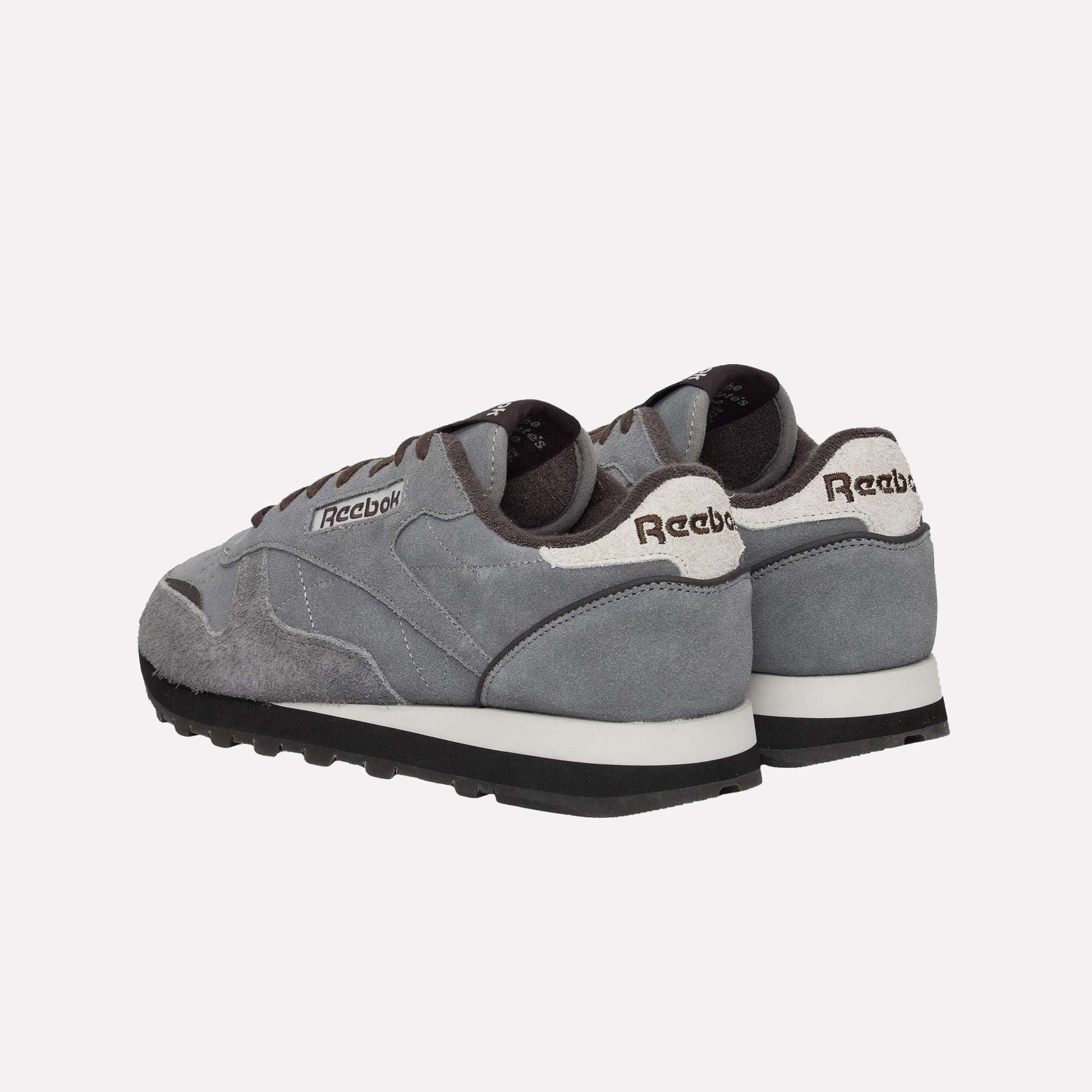 A pair of Reebok Classic Leather Premium LTD Shoes in gray, featuring white soles and black bottoms, are displayed on a light blue background with back and side views that highlight their retro, timeless silhouette.