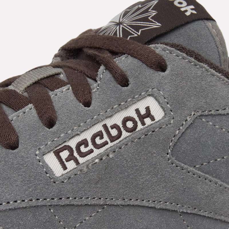 Close-up of a grey suede Reebok Classic Leather Premium LTD Shoe with brown laces. The Reebok logo and part of a star logo are visible, highlighting its classic silhouette against a light blue background.
