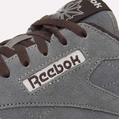 Close-up of a grey suede Reebok Classic Leather Premium LTD Shoe with brown laces. The Reebok logo and part of a star logo are visible, highlighting its classic silhouette against a light blue background.