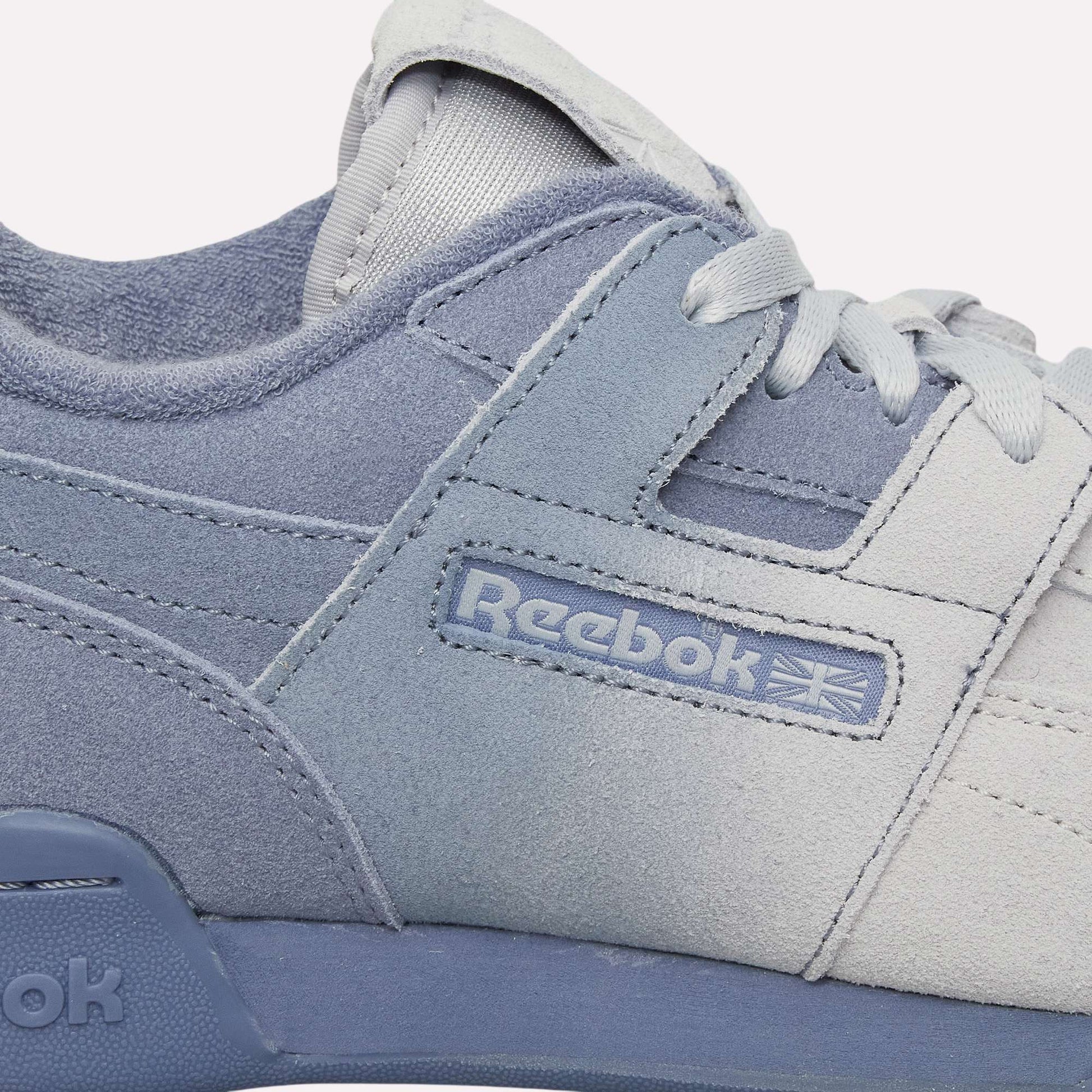 Close-up of light blue and white Reebok Workout Plus Og Shoes with visible side logo, laces, and stitching. The classic retro silhouette stands out against a light blue background.