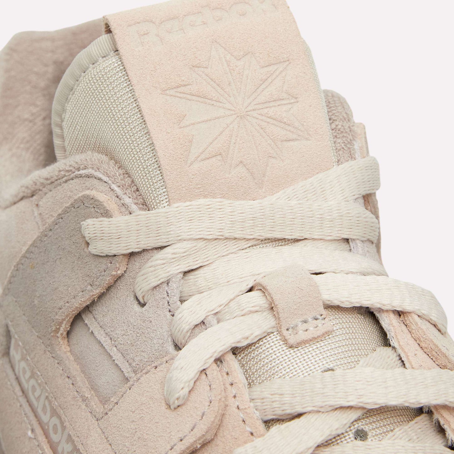Close-up of beige Reebok Workout Plus Og Shoes highlights textured suede, off-white laces, and an embossed Reebok logo with a star on the tongue. These retro sneakers feature a classic silhouette set against a light blue background.