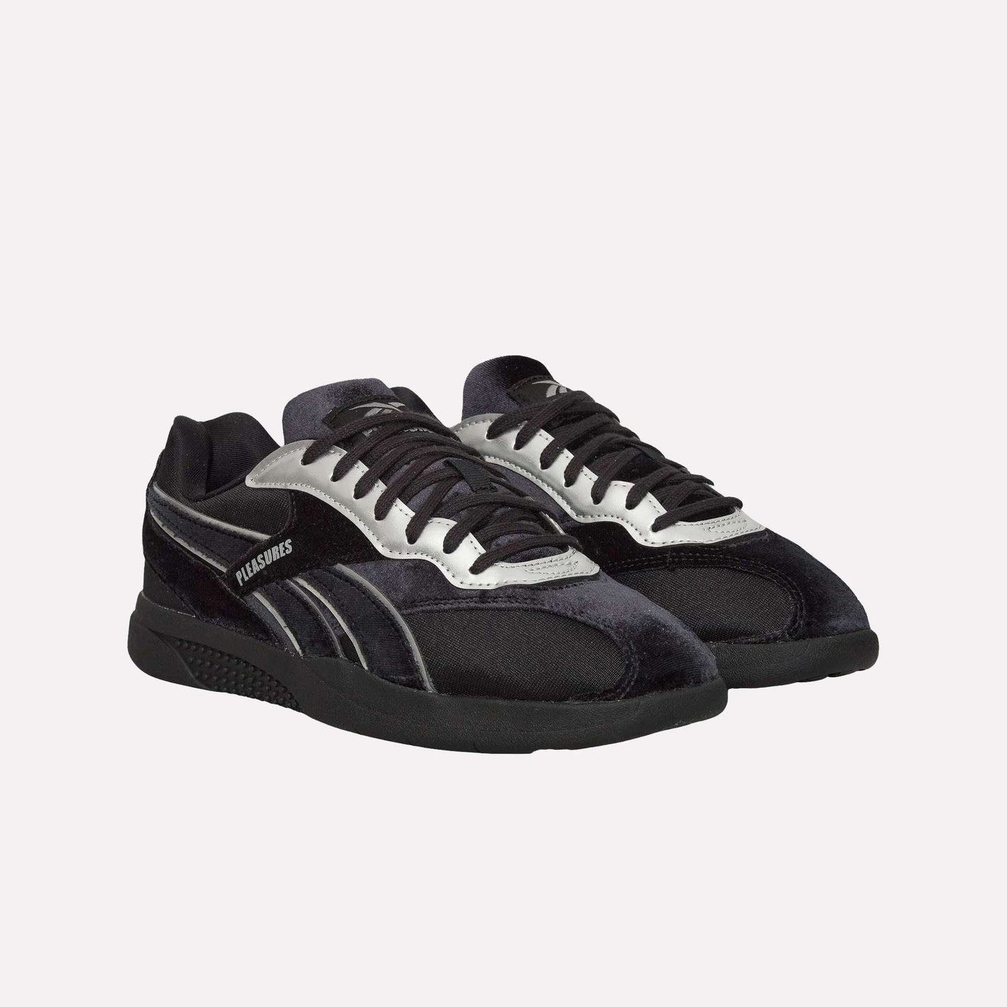 The Reebok Pleasures Hammer Street Shoes are black and silver low-top sneakers with black laces and PLEASURES branding, inspired by classic Reebok indoor soccer styles, shown on a light blue background.