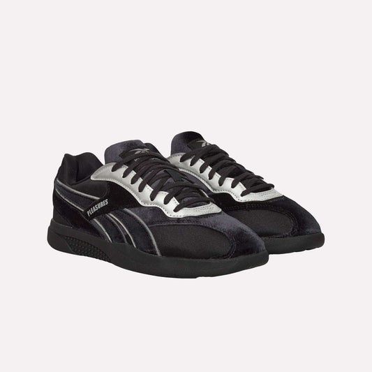 The Reebok Pleasures Hammer Street Shoes are black and silver low-top sneakers with black laces and PLEASURES branding, inspired by classic Reebok indoor soccer styles, shown on a light blue background.
