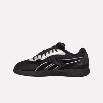 The Reebok Pleasures Hammer Street Shoes feature a black and white low-top design with black laces, silver accents, and a velvet upper inspired by Reebok indoor soccer shoes, shown against a plain light blue background.