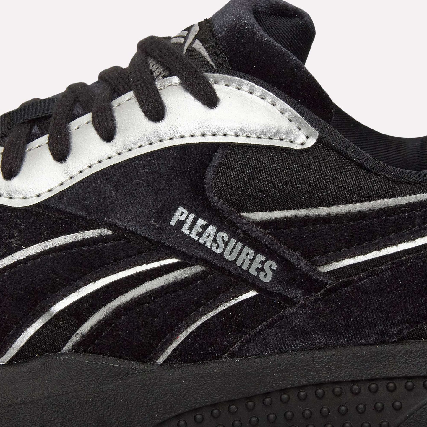 Close-up of Pleasures Hammer Street Shoes by Reebok, a black sneaker with white accents inspired by indoor soccer styles, "PLEASURES" printed on the side panel, textured material, black laces, and featuring the PLEASURES collaboration.