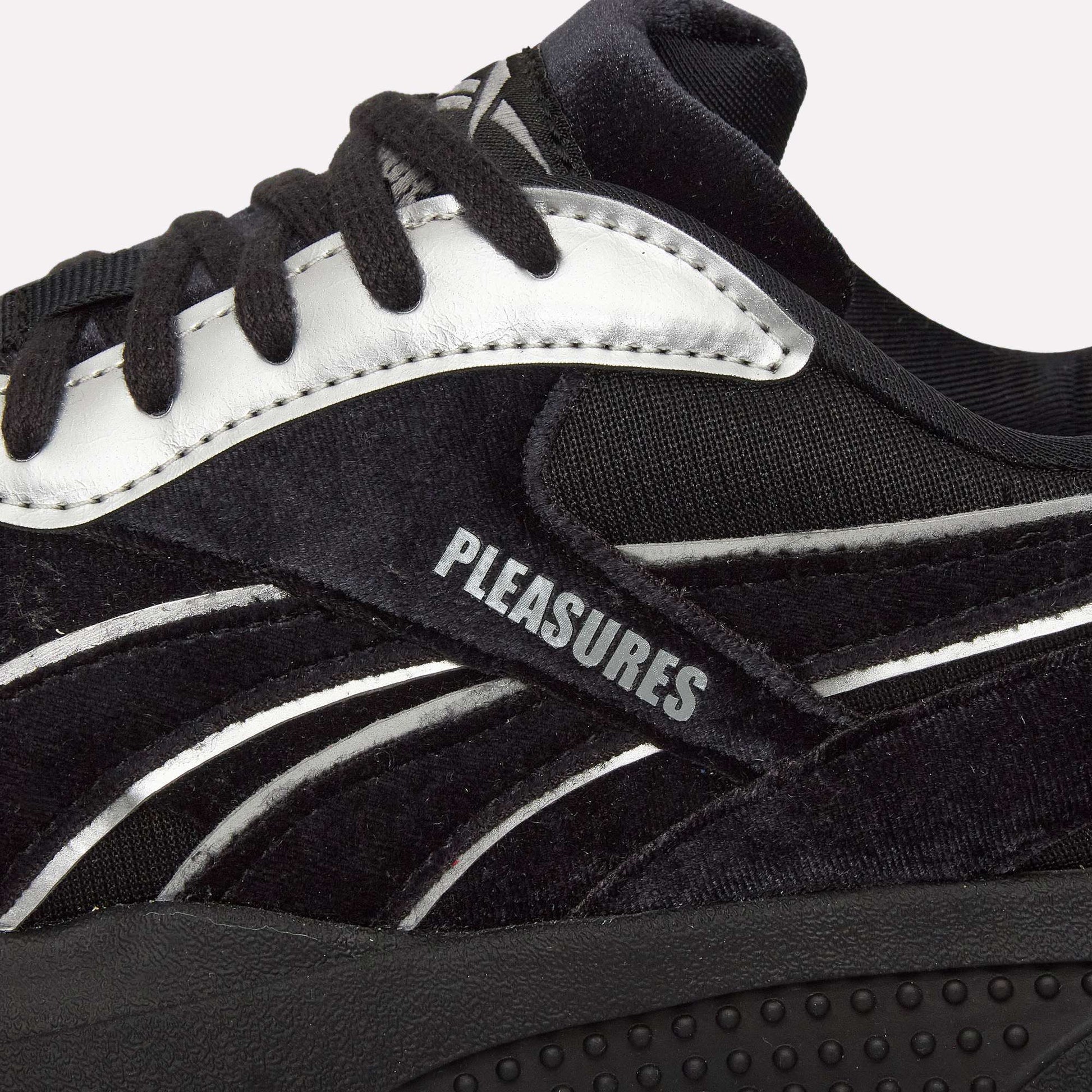 Close-up of Pleasures Hammer Street Shoes by Reebok, a black sneaker with white accents inspired by indoor soccer styles, "PLEASURES" printed on the side panel, textured material, black laces, and featuring the PLEASURES collaboration.