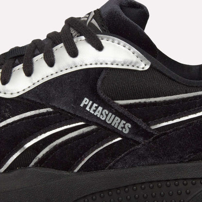 Close-up of Pleasures Hammer Street Shoes by Reebok, a black sneaker with white accents inspired by indoor soccer styles, "PLEASURES" printed on the side panel, textured material, black laces, and featuring the PLEASURES collaboration.
