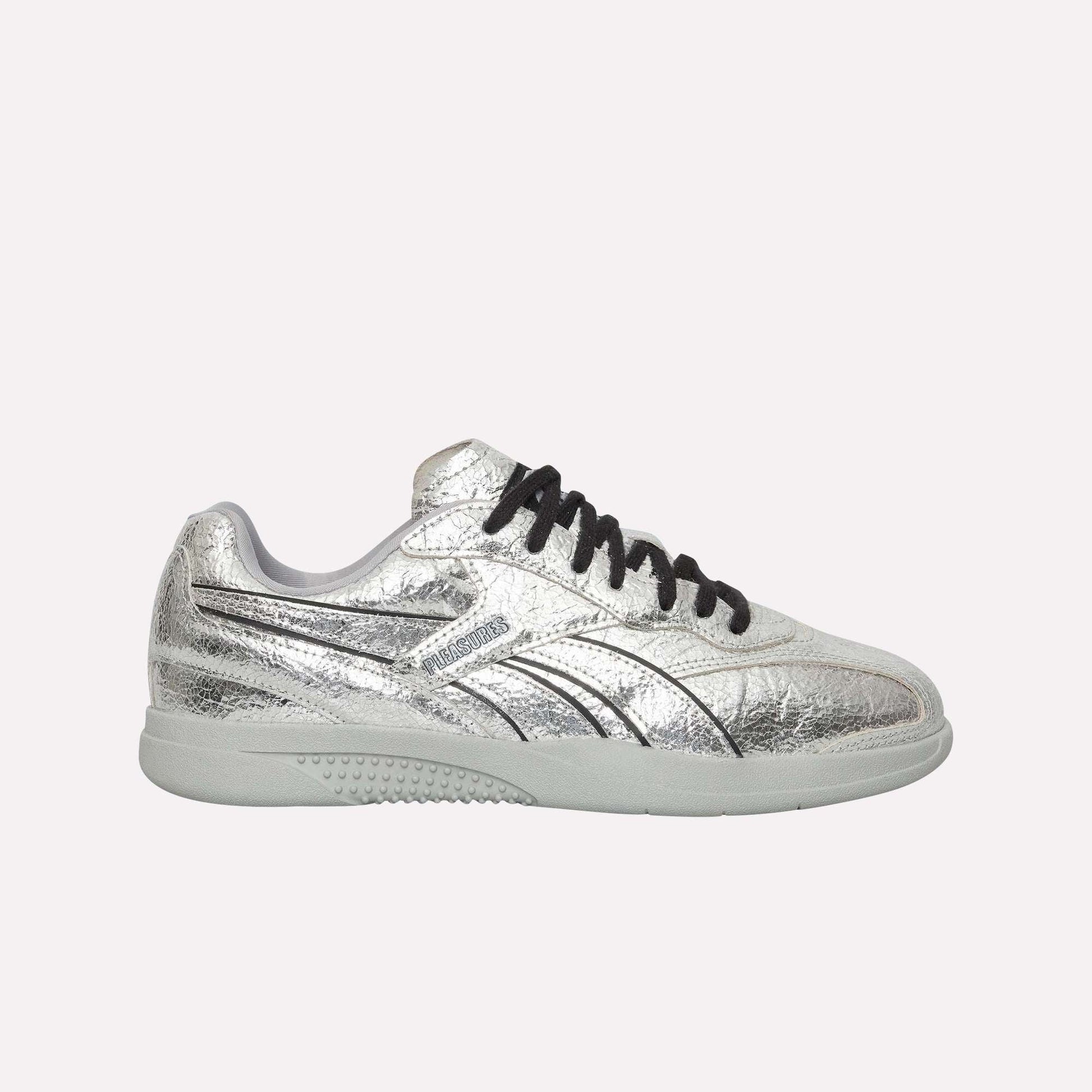 The Reebok Pleasures Hammer Street Shoes, shown in silver leather with black laces, a white sole, black line accents, and a subtle textured finish, are inspired by classic Reebok indoor soccer shoes against a light blue background.