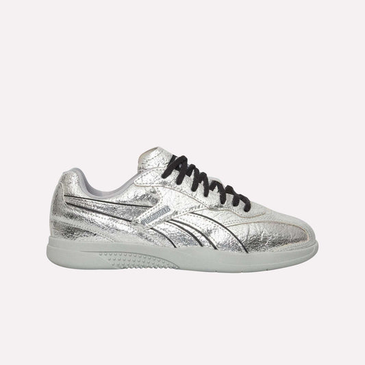 The Reebok Pleasures Hammer Street Shoes, shown in silver leather with black laces, a white sole, black line accents, and a subtle textured finish, are inspired by classic Reebok indoor soccer shoes against a light blue background.