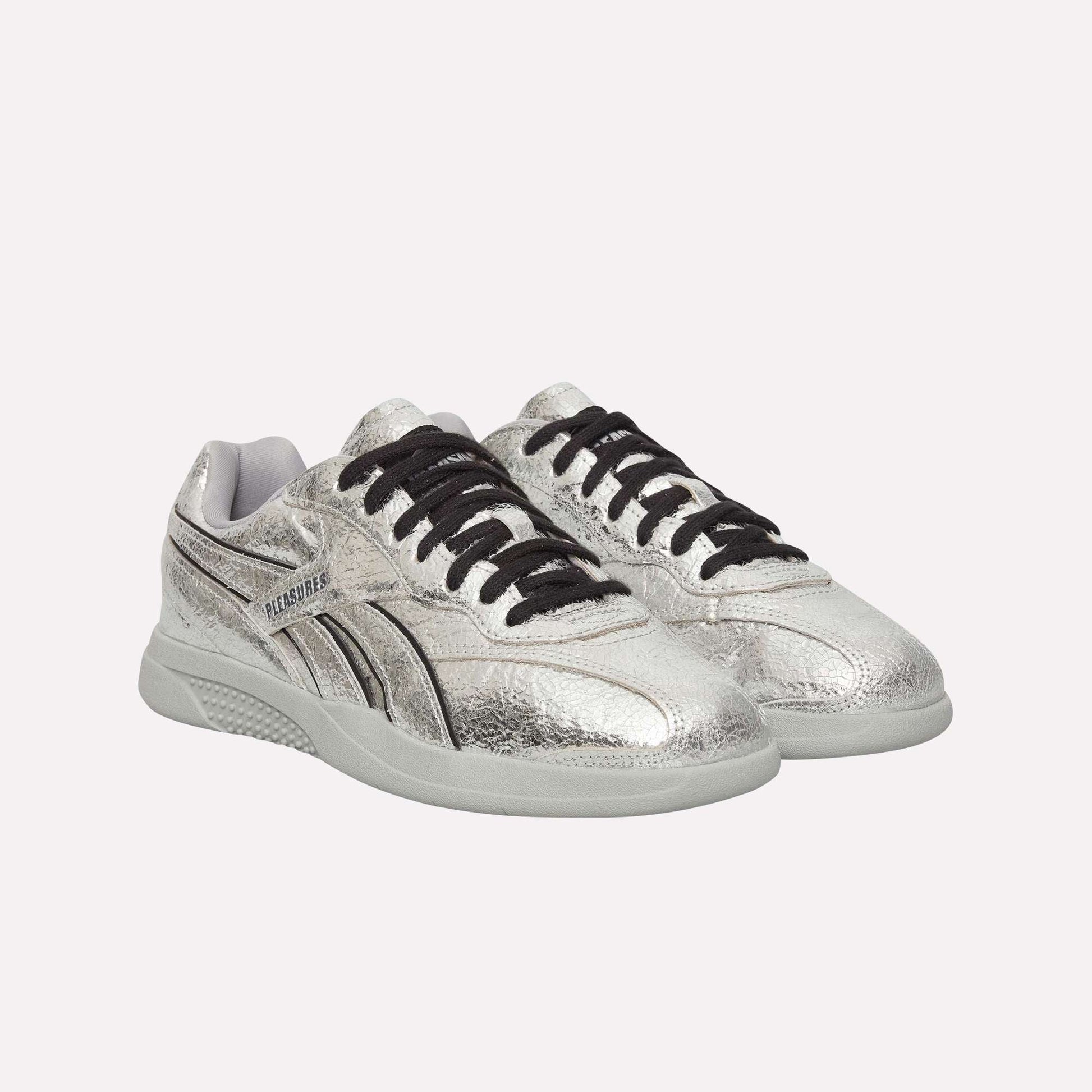 The Pleasures Hammer Street Shoes by Reebok are silver leather sneakers with black laces and white soles, featuring a textured finish and side logo detail, inspired by classic Reebok indoor soccer styles. Displayed on a light blue background.