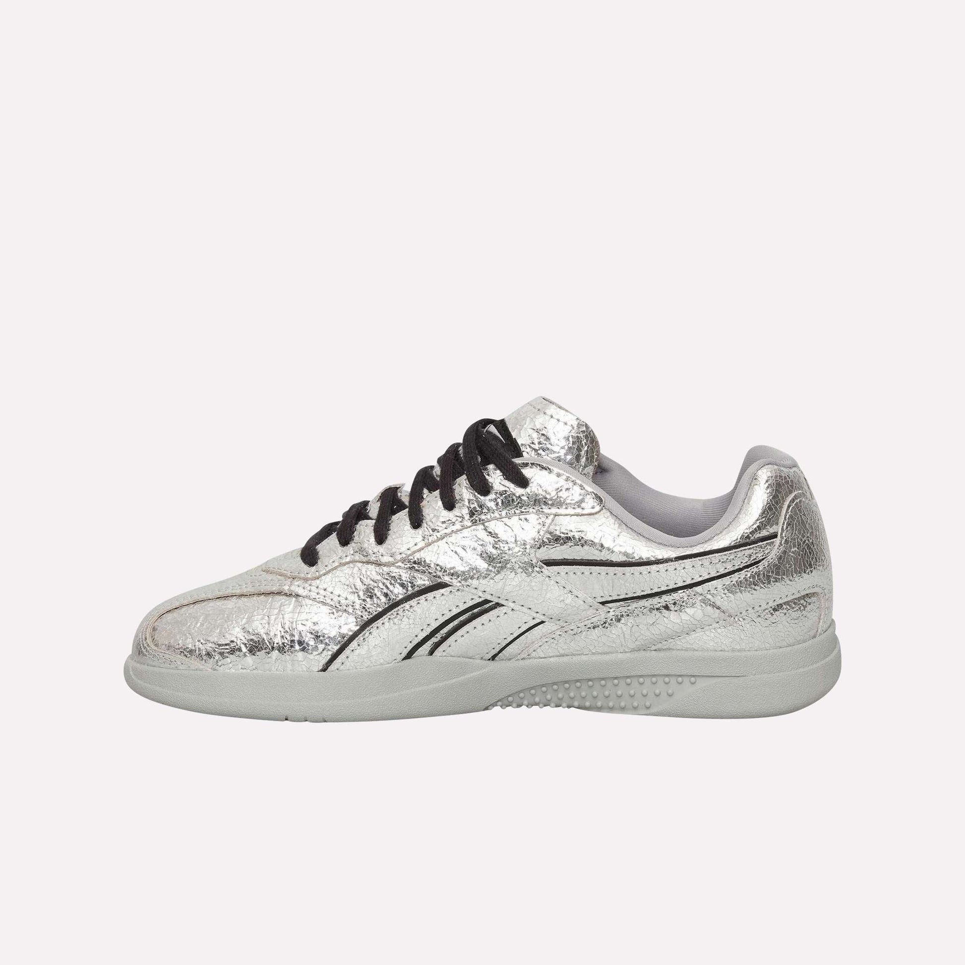 The Reebok Pleasures Hammer Street Shoes, featuring shiny silver leather, black laces, and swooping design details, are displayed from the side on a light blue background.