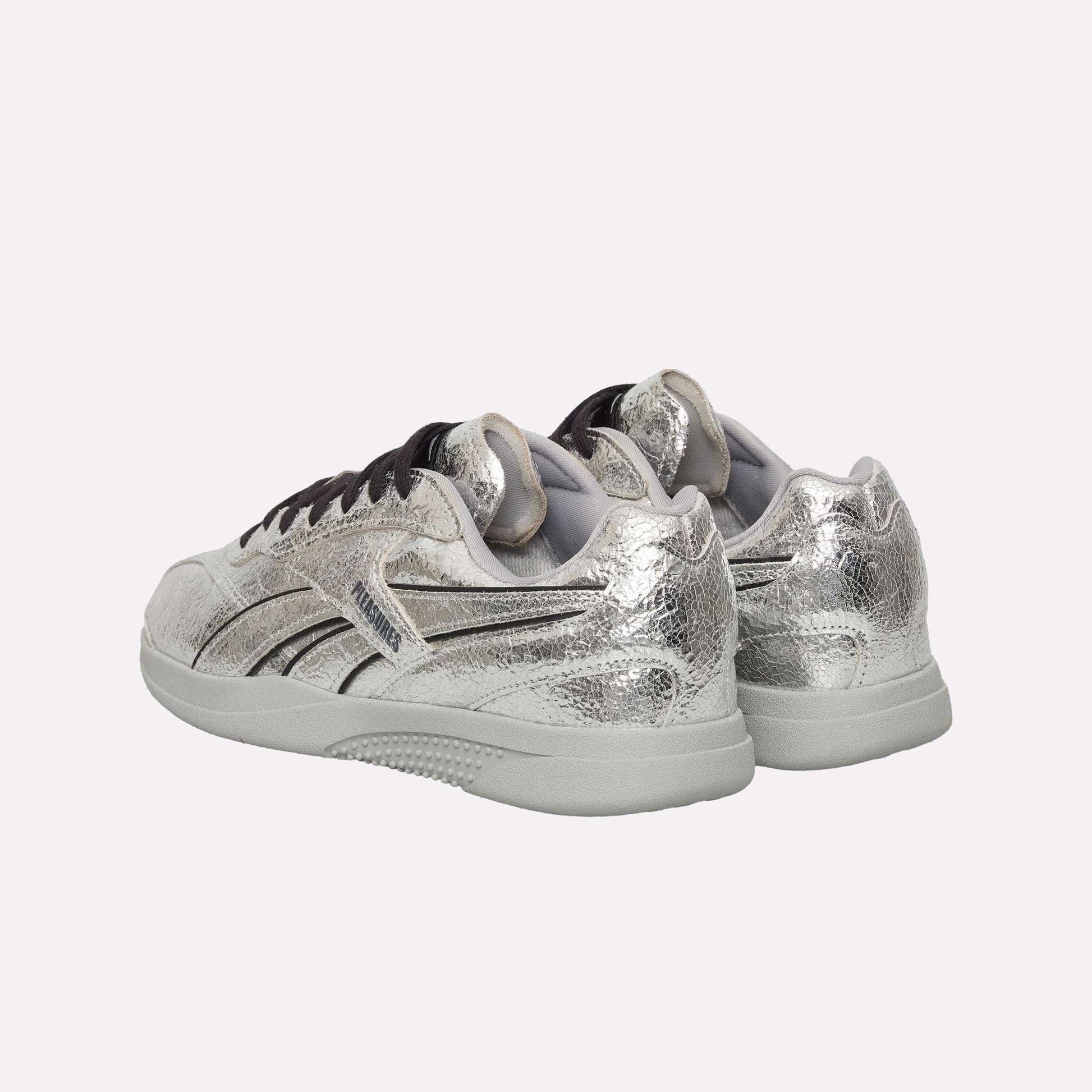 A pair of Reebok Pleasures Hammer Street Shoes in metallic silver leather with black stripes and accents, shown from the back angled left, on a light blue background—part of a PLEASURES collaboration.