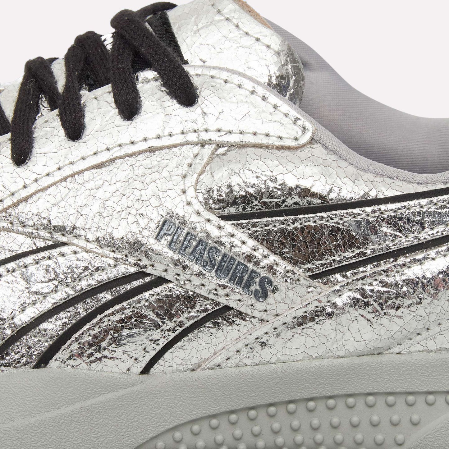 Close-up of Reebok Pleasures Hammer Street Shoes in silver leather with a cracked texture, black laces, light gray inner lining, and Pleasures branding on the side above a textured light gray sole.