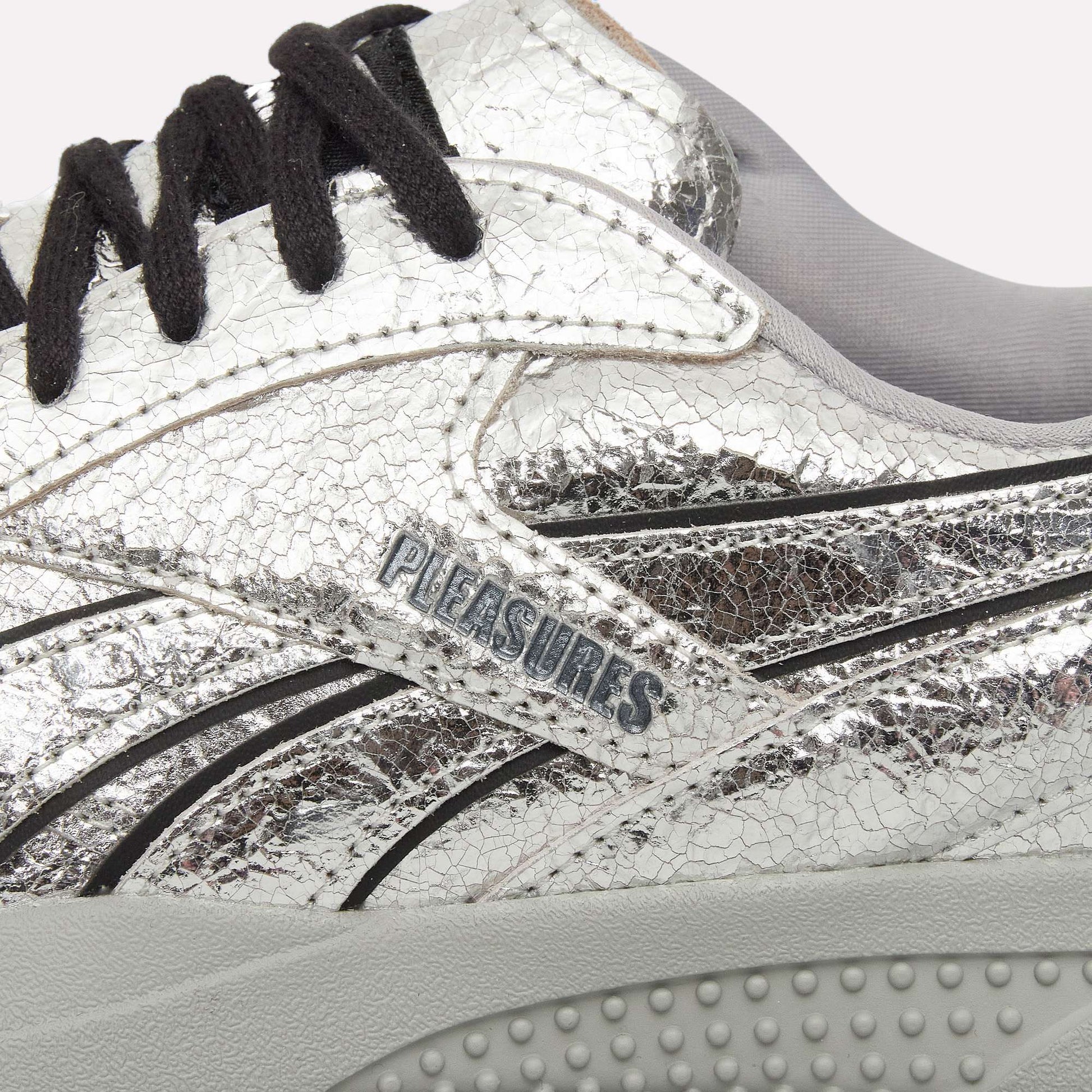 Close-up of Reebok Pleasures Hammer Street Shoes in silver leather with a cracked texture, black laces, light gray inner lining, and Pleasures branding on the side above a textured light gray sole.