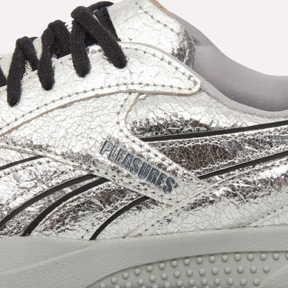 Close-up of Reebok Pleasures Hammer Street Shoes in silver leather with a cracked texture, black laces, light gray inner lining, and Pleasures branding on the side above a textured light gray sole.