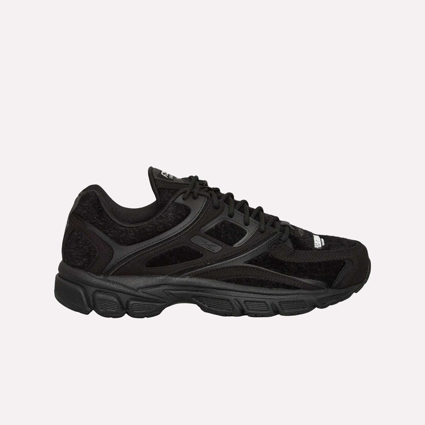 The Reebok Pleasures Premier Trinity KFS Shoes feature a black athletic design with mesh and suede details, black laces, a textured rubber sole, and subtle Reebok branding. Displayed on a light blue background.