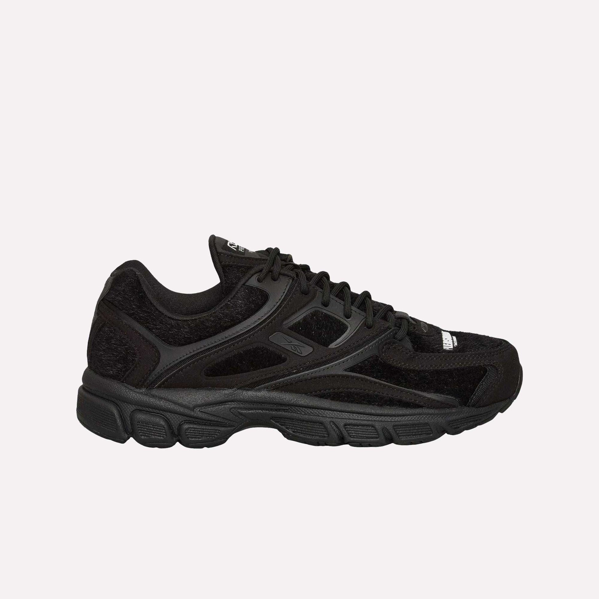 The Reebok Pleasures Premier Trinity KFS Shoes feature a black athletic design with mesh and suede details, black laces, a textured rubber sole, and subtle Reebok branding. Displayed on a light blue background.