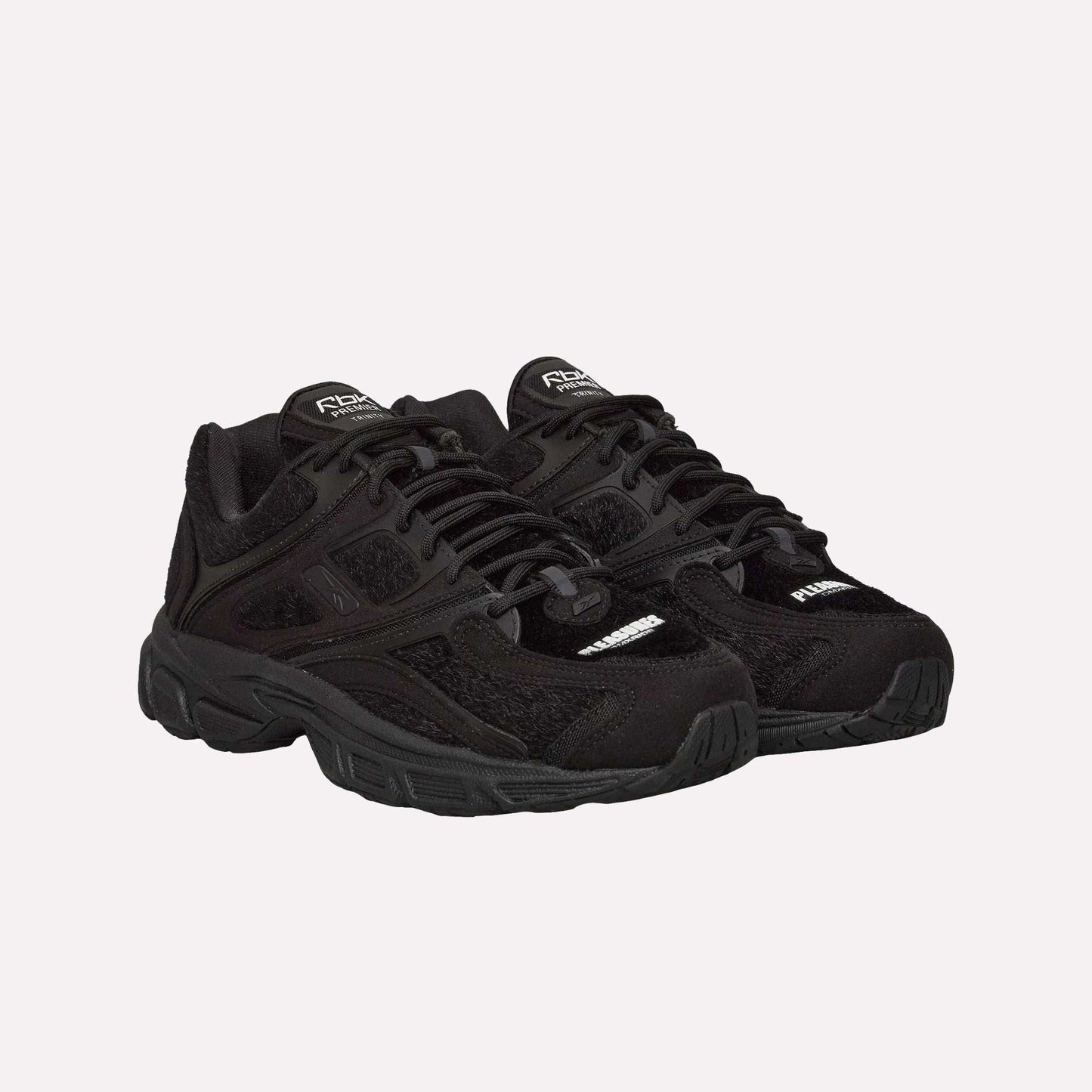 The Reebok Pleasures Premier Trinity KFS Shoes are black athletic sneakers with thick soles, mesh accents, and a unique pony hair upper, displayed on a plain light blue background.
