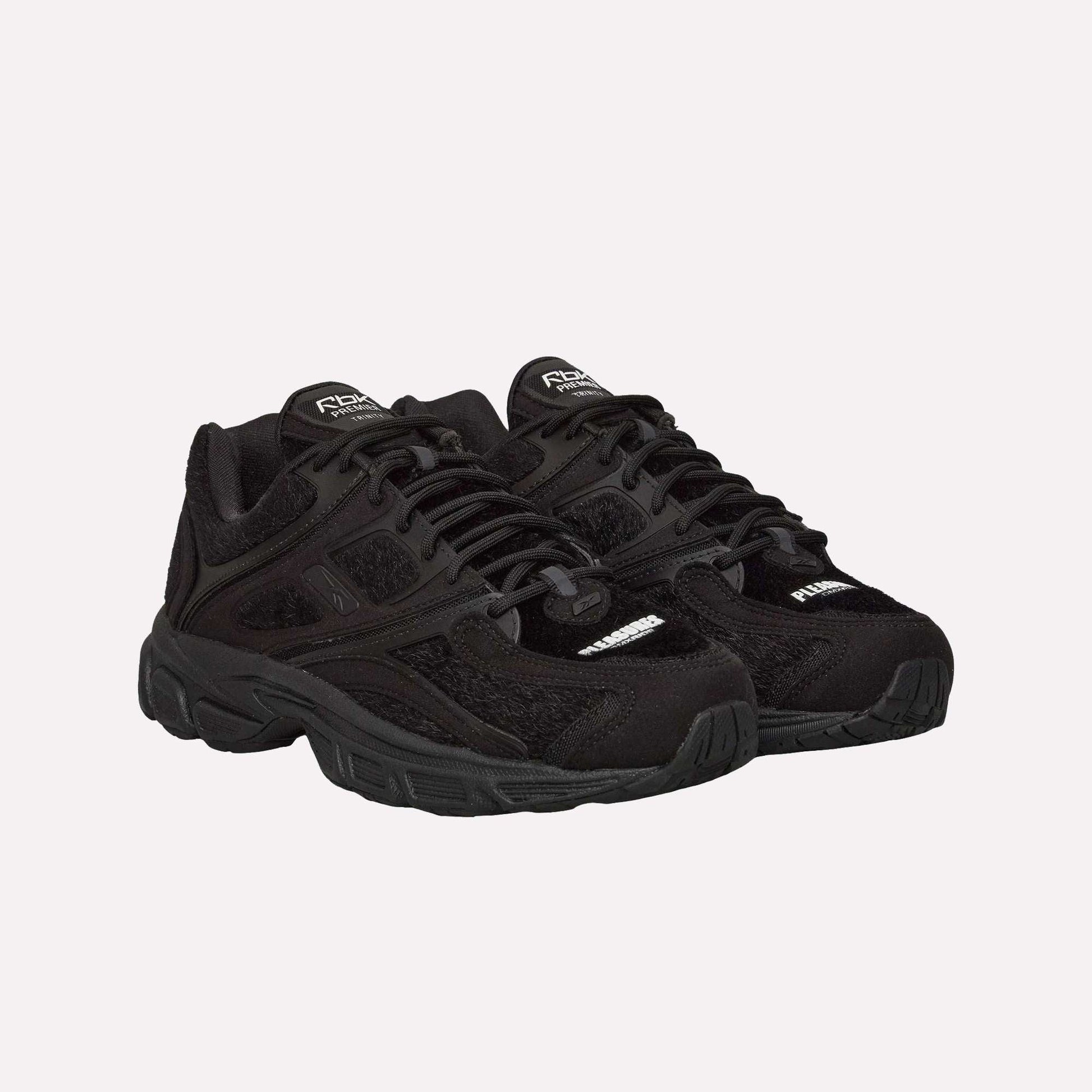 The Reebok Pleasures Premier Trinity KFS Shoes are black athletic sneakers with thick soles, mesh accents, and a unique pony hair upper, displayed on a plain light blue background.
