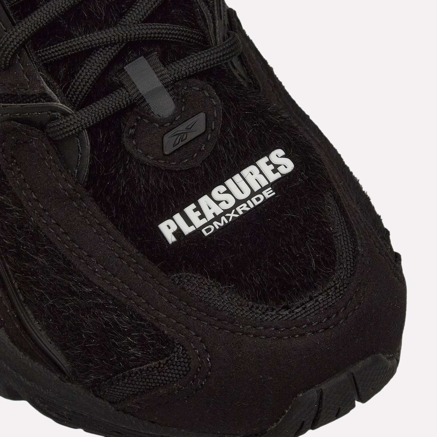 Close-up of the Pleasures Premier Trinity KFS Shoes from Reebok, featuring a black textured pony hair upper. "PLEASURES" stands out in bold white with DMXRIDE underneath. Finished with black laces and stitching for a striking look.
