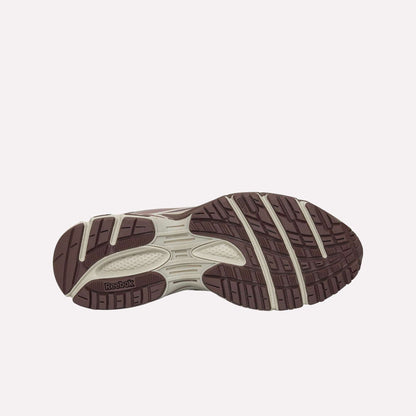 The image displays the outsole of the Reebok Premier Road Modern, showcasing a brown and beige pattern with textured grips for traction, a dual-density midsole for comfort, and the Reebok logo on the sole.