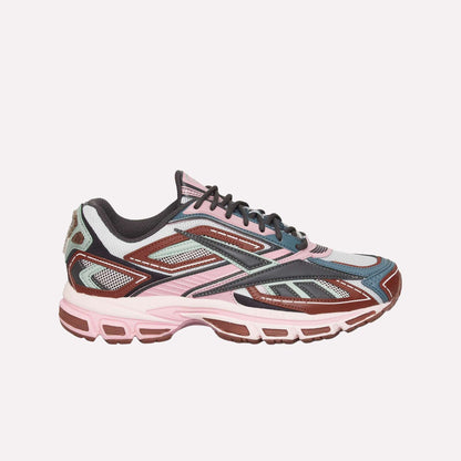 The Reebok Premier Road Ultra is a stylish sneaker in pink, maroon, gray, and black with mesh panels, detailed overlays, and a dual-density midsole—shown in side profile against a white background.