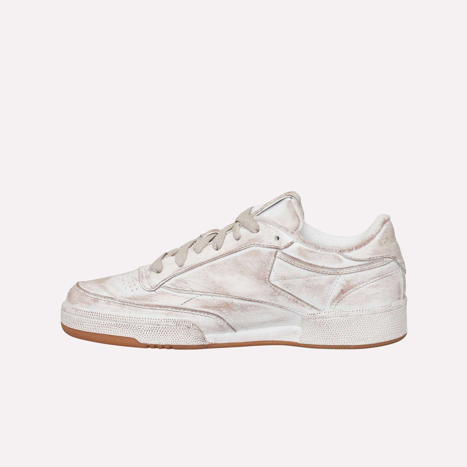 A side view of the Reebok Club C 85 Vintage, a low-top sneaker with a worn white finish, gray laces, textured leather details, and a brown rubber sole. The distressed look adds timeless appeal to this classic tennis-inspired design.