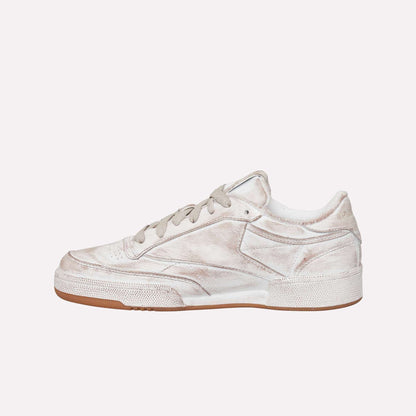 A side view of the Reebok Club C 85 Vintage, a low-top sneaker with a worn white finish, gray laces, textured leather details, and a brown rubber sole. The distressed look adds timeless appeal to this classic tennis-inspired design.