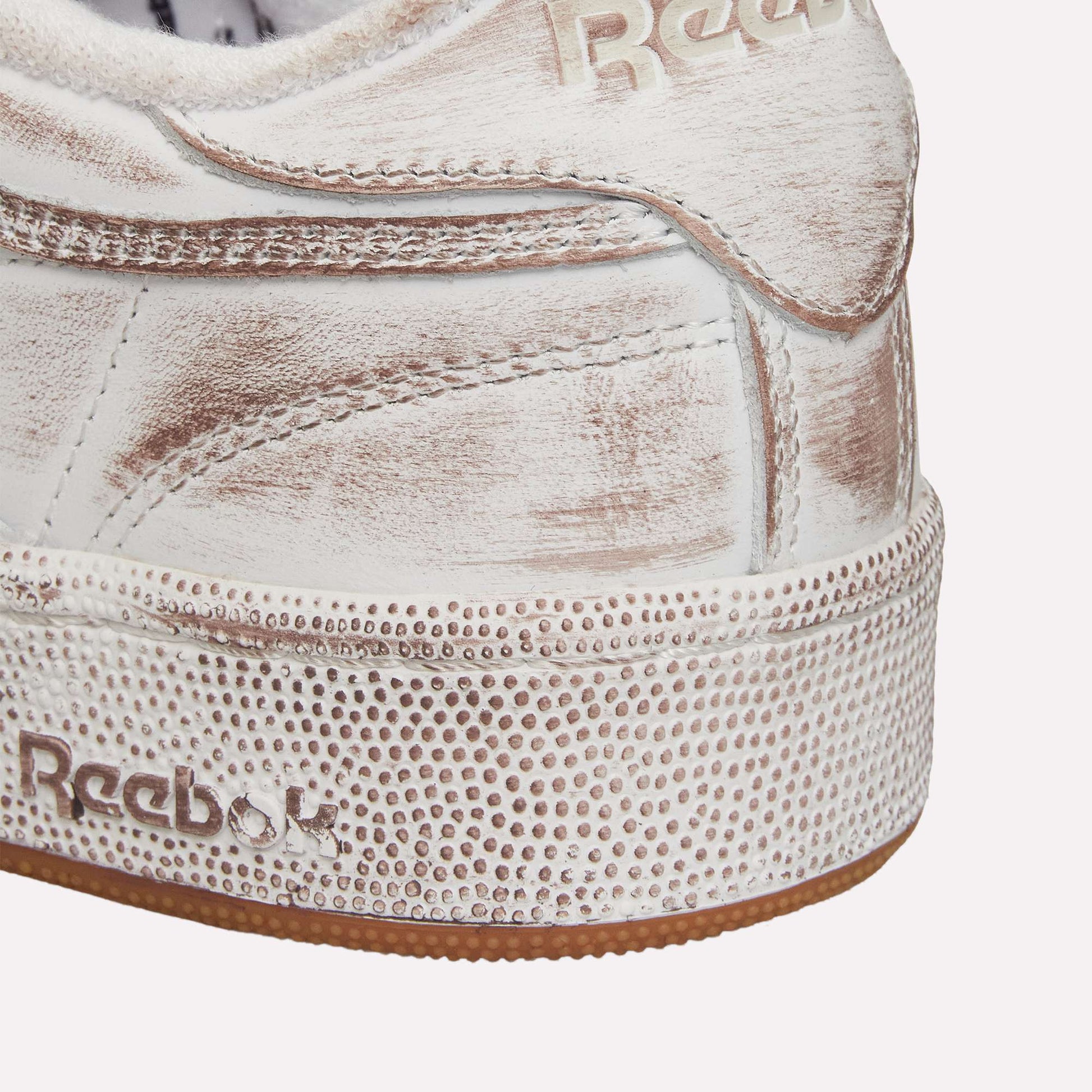 Close-up of the heel of a white Reebok Club C 85 Vintage tennis shoe with distressed leather and midsole, textured dot detailing, and embossed Reebok logos—a classic silhouette with updated vintage style.